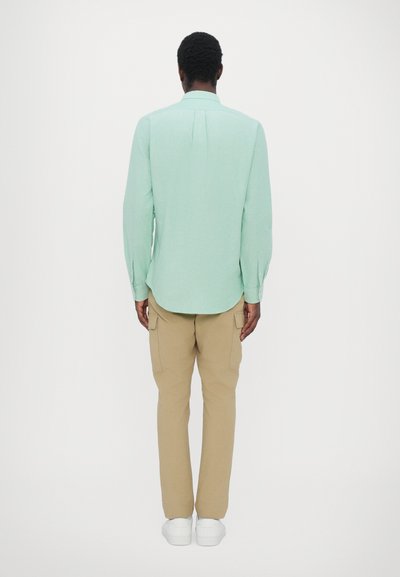 Chemise à manches longues vert menthe avec une coupe droite et un pli dans le dos. Associée à un pantalon cargo beige et des baskets blanches, vue de dos.