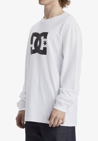 White long-sleeve t-shirt made of cotton, featuring a large black DC logo with a star on the front. Loose fit with round neckline and cuffed sleeves.