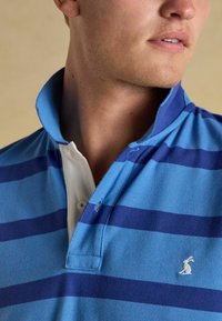 Blaues Polohemd mit vertikalen marineblauen Streifen, weißem Kragen, einer Dreiknopfleiste und einem kleinen weißen Hasenlogo auf der Brust.