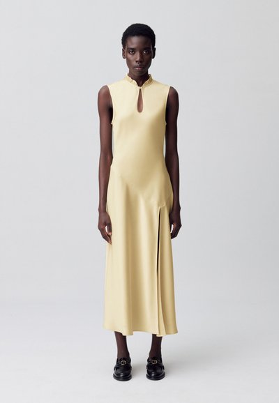 Abito senza maniche, lungo fino al ginocchio, in morbido satin giallo chiaro. Caratterizzato da un collo a chiave e una fessura laterale per la libertà di movimento. Decorato con un colletto arricciato.