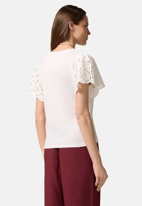Witte top met korte mouwen en kantaccenten op de mouwen, zachte textuur, ronde halslijn, gecombineerd met bordeauxrode broek.
