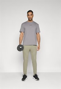 Tricou gri cu mânecă scurtă, pantaloni scurți tip jogger verzi măslinii, și încălțăminte sportivă neagră; ținând o placă de greutate neagră în mâna dreaptă.