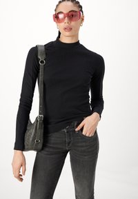 Nainen, jolla on letitetyt hiukset, pukeutuneena suuriin vaaleanpunaisiin aurinkolaseihin, mustaan pitkähihaiseen paitaan, tummiin farkkuihin ja olkalaukkuun, poseeraa valkoista taustaa vasten.