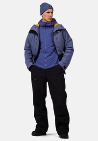 Robuste Outdoor-Jacke in Blau mit gelben Akzenten, kombiniert mit schwarzen wasserdichten Hosen. Verfügt über Reißverschlusstaschen und eine gemütliche Mütze.