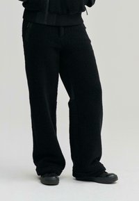 Fekete, bolyhos fleece nadrág, laza szabással, oldalsó zsebekkel és puha textúrával, fekete, lapos talpú cipőkkel párosítva.