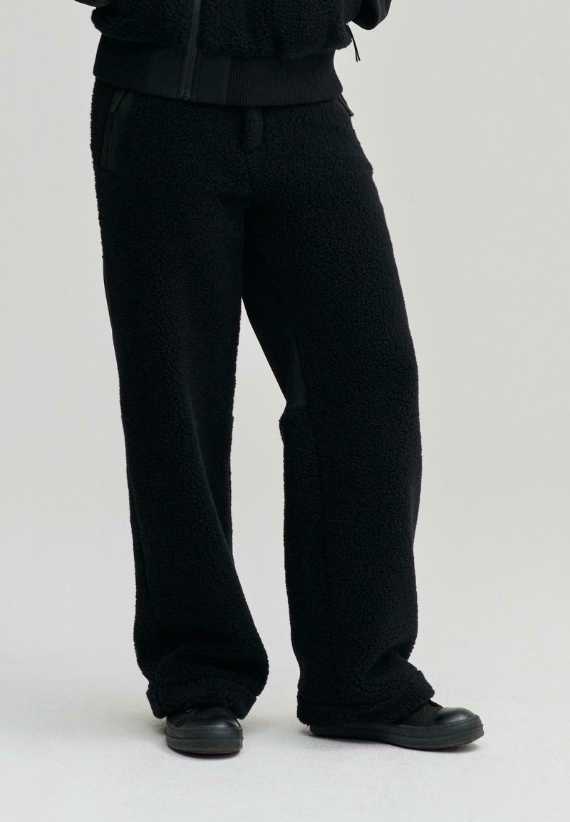 Fekete, bolyhos fleece nadrág, laza szabással, oldalsó zsebekkel és puha textúrával, fekete, lapos talpú cipőkkel párosítva.