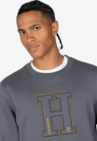Szary sweatshirt z dużą haftowaną literą blokową "H" w ciemnoniebieskim i żółtym haftem, z napisem "HARPER" w żółtej czcionce poniżej.