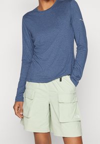 Osoba oblečená v modré košili s dlouhým rukávem a světlých zelených cargo šortkách s velkými předními kapsami, stojící s jednou rukou v kapse.