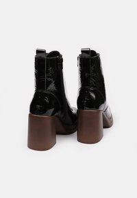 Botas de tobillo de charol negro con un alto tacón rectangular, cierre de cremallera lateral y textura suave. Presenta un minimalismo en los detalles.