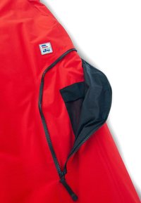 Jachetă roșie cu un buzunar cu fermoar, căptușit cu plasă neagră, bandă de fermoar neagră și un patch logo pe umăr. Material sintetic neted.