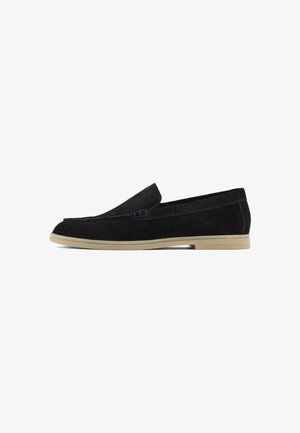 Mocassins en cuir suédé noir avec une semelle plate beige, présentant un design cousu et un style slip-on élégant, adaptés pour un usage décontracté.