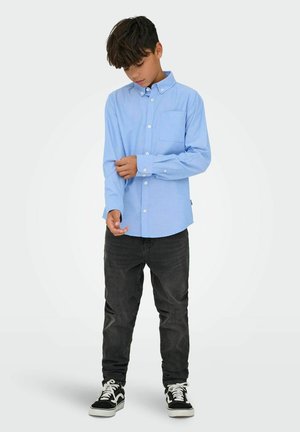 Boy wearing a light blue button-up shirt, black jeans, and black sneakers, adjusting his left shirt sleeve, standing on a plain background.