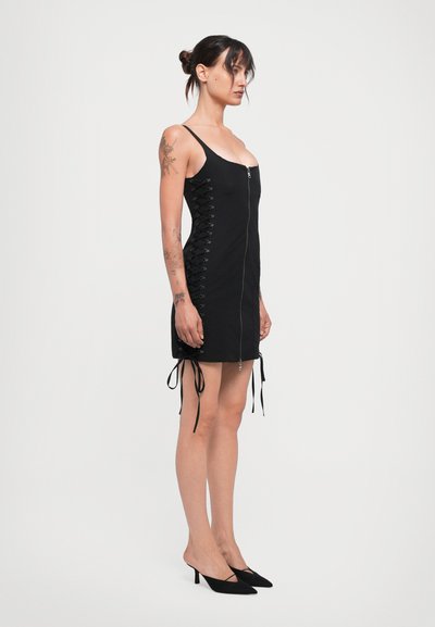 Black sleeveless dress with a front zipper, lace-up sides, and side drawstrings. Smooth fabric, knee-length, paired with black pointed heels.