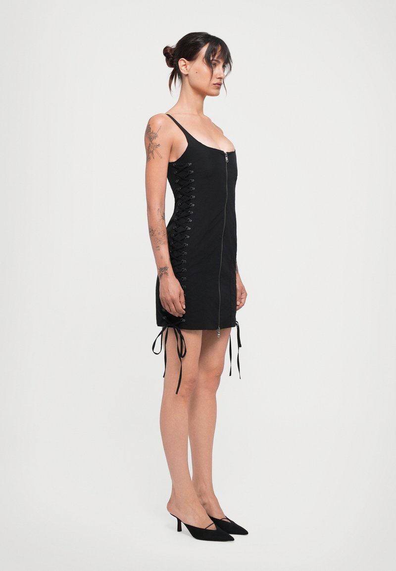 Svart ärmlös klänning med en framdragkedja, snörning i sidorna och dragsko på sidorna. Slätt tyg, knälång, kombinerad med svarta spetsiga klackar.