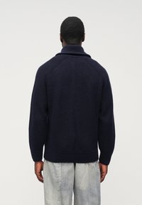 Tmavomodrý pletený sveter s klopovým golierom, rebrovanou textúrou a spustenými ramenami, v kombinácii so svetlosivými nohavicami.