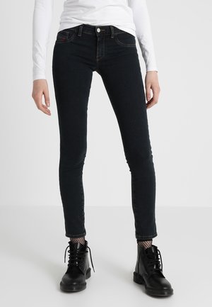 Henkilö, jolla on mustat kapeat farkut, mustat nauhalliset saappaat, kalaverkkosukat ja valkoinen pitkähihainen paita, seisoo vaaleanharmaalla lattialla.