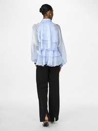 Himmelblaues Seidenblusenshirt mit geschichteten Rüschen auf der Rückseite; kombiniert mit schwarzen, taillierten Hosen mit Schlitzsäumen und Stilettos.
