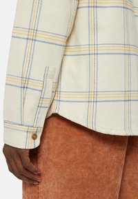 Camisa de franela a cuadros en crema, azul y amarillo con puños abotonados y tejido texturizado; combinada con pantalones de pana color óxido.