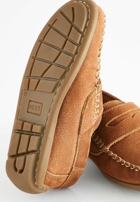 Des chaussures en cuir marron à enfiler présentent un dessus texturé et des coutures décoratives, avec une semelle en caoutchouc affichant un motif de grille pour l'adhérence.