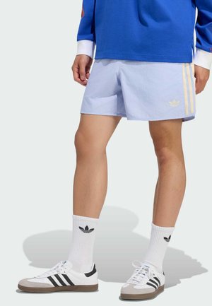 Mand iført lyseblå Adidas-shorts med beige striber, hvide Adidas-strømper og hvide sneakers med sorte striber, der står på en hvid baggrund.