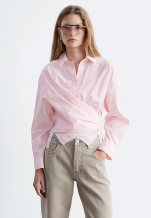 Jeune femme portant des lunettes aviateur transparentes, une chemise cache-cœur rose clair et un pantalon beige sur fond blanc uni.