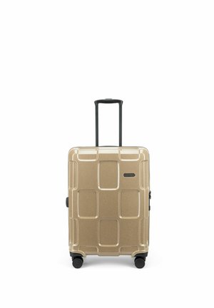 Valise rigide dorée avec quatre roues noires pivotantes et une poignée télescopique noire déployée en position verticale.