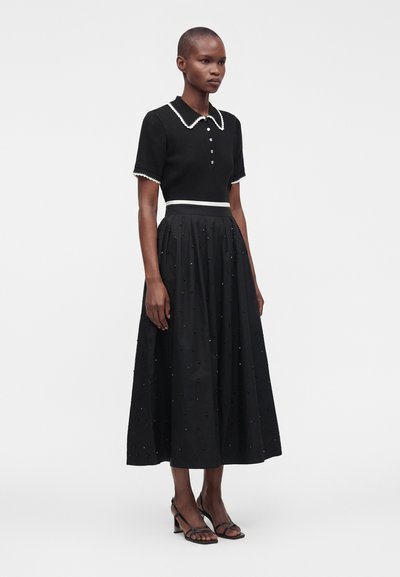 Femme portant un haut noir à manches courtes avec col et bordure blanche et une jupe noire longue jusqu'aux chevilles avec de subtils ornements en perles.
