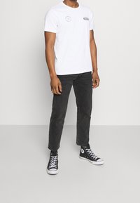 White cotton t-shirt with printed logos on the chest, paired with black denim jeans and black high-top sneakers with white soles and laces.