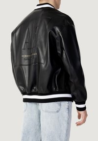 Svart läderbomberjacka med vita och svarta randiga ribbade muddar. Har präglingen "AX"-logotyp och guldtext på ryggen. Avslappnad passform.