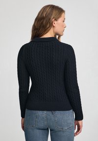 Kobieta stojąca bokiem, ubrana w granatowy sweter w warkocze oraz niebieskie dżinsy, na tle gładkiego jasnoszarego tła.