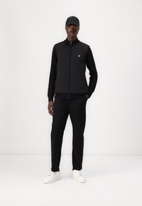 Black quilted vest over a black sweater, paired with black pants and white sneakers. A yellow logo is visible on the vest. Black cap worn.