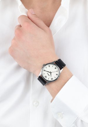 Silberne Uhr mit rundem Zifferblatt, weiß gesprengtem Ziffernblatt, schwarzem Lederarmband und sichtbaren Stundenmarkierungen. Am Handgelenk über einem weißen Hemd getragen.