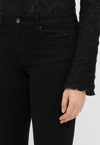 Melnas augsta vidukļa džinsos ar šauru piegriezumu, izgatavoti no denima. Iekļauj kabatas, pogu aizdari un teksturētas mežģīņu piedurknes detaļas.