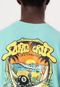 T-shirt de algodão turquesa com uma impressão gráfica apresentando uma cena de praia, palmeiras, uma bola 8 e o texto "Santa Cruz Skateboards" em amarelo.