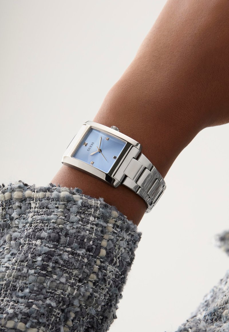 Montre en métal argenté avec un cadran rectangulaire, cadran bleu clair, quatre index horaires métalliques et un bracelet à maillons, portée sur un poignet avec un tissu texturé.