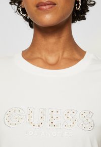 Camiseta de algodón blanca con la inscripción "GUESS LOS ÁNGELES" en un diseño texturizado y perforado con acentos en plata y cuello redondo.