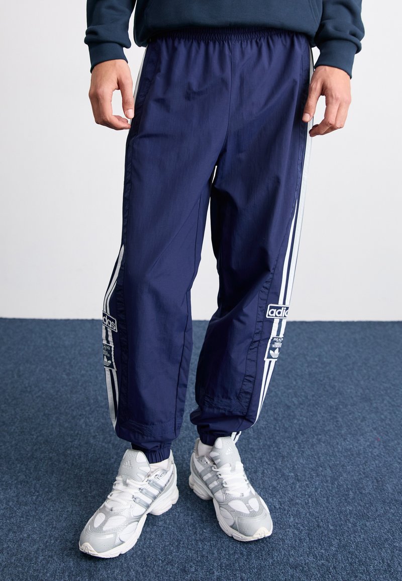 Navy träningsoveraller med vita sidostripes och elastiska mudd, med en adidas-logotyp och en strömlinjeformad design, tillverkade av lättviktsmaterial.