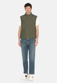 Gilet senza maniche verde oliva con colletto alto, zip frontale e orlo elasticizzato, abbinato a una maglietta bianca e jeans blu. Comprende anche sneakers bianche.