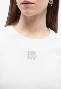 Νεαρός με ίσια καστανά μαλλιά που φορά ασημένια σκουλαρίκια κρίκους και λευκό πουκάμισο με λογότυπο DKNY σε ρόμβους στρας στο στήθος.