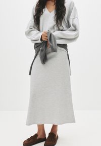 Haut gris à manches longues, col en V avec coupe décontractée ; jupe grise assortie ; texture douce ; vêtement gris noué à la taille ; mocassins marron.