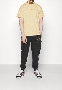 Person med vitiligo iklädd en beige Nike t-shirt, svarta Nike cargo-byxor och sneakers i vitt, svart och rött stående mot en vit bakgrund.