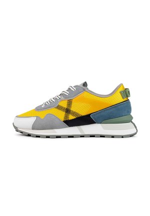 Zapatilla gris y amarilla con materiales de malla y ante. Presenta un panel texturizado amarillo, acentos negros y una lengüeta en el talón de color verde. Suela de goma.