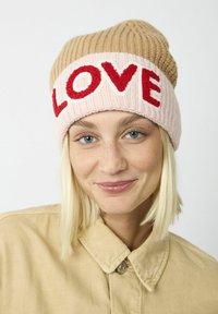 Gehaakte beanie in beige en roze, met "LOVE" in rode garen. Ribbelstructuur, slouchy ontwerp en opvallende contrasterende kleuren.
