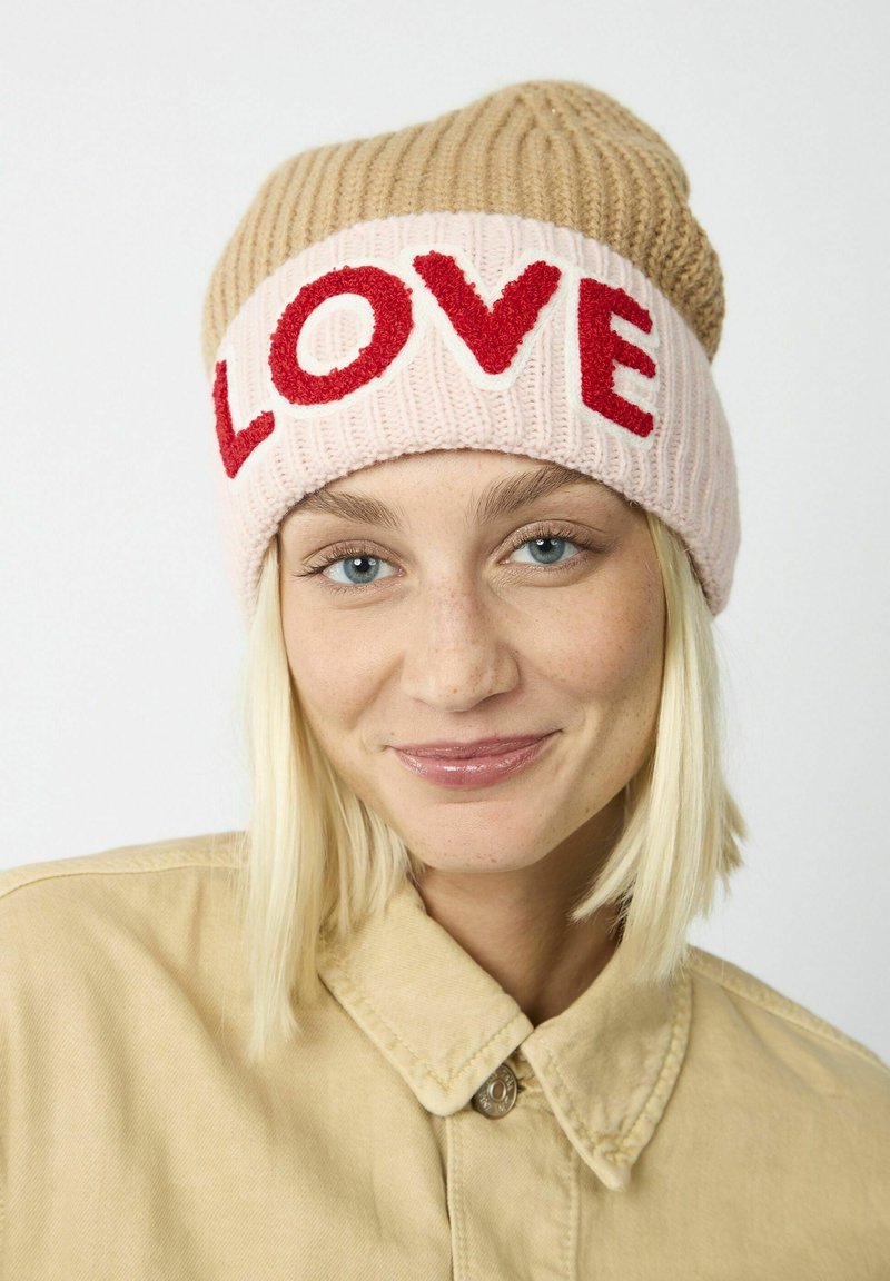Gehaakte beanie in beige en roze, met "LOVE" in rode garen. Ribbelstructuur, slouchy ontwerp en opvallende contrasterende kleuren.