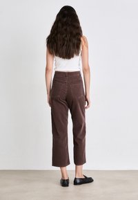 Bruine corduroy hoge taille broek met een rechtonder ontwerp en zichtbare achterzakken. Het model draagt een witte tanktop en zwarte schoenen.