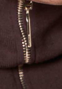 Brązowy, zapinany na zamek ciemny element garderoby o gładkiej fakturze. Zamek błyskawiczny w kolorze złotym z widocznymi zębami i suwakiem, podkreślający szczegóły jego designu.