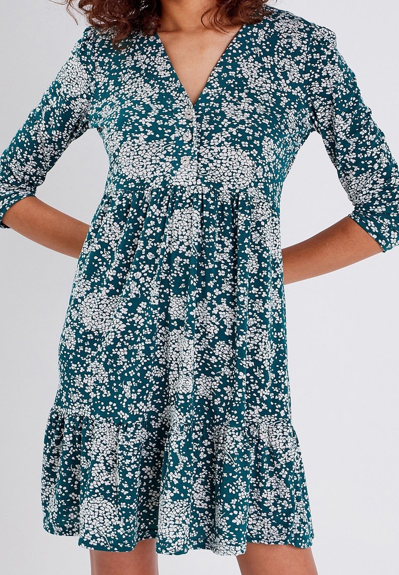 Femme portant une robe turquoise à motifs floraux blancs et manches trois-quarts, mains posées sur les hanches, debout devant un fond uni.
