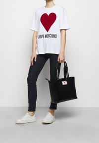 Weißes T-Shirt mit einem roten Herz-Design und dem Text "LOVE MOSCHINO", kombiniert mit dunklen Jeans und einer eleganten schwarzen Lackhandtasche. Weiße Sneakers.
