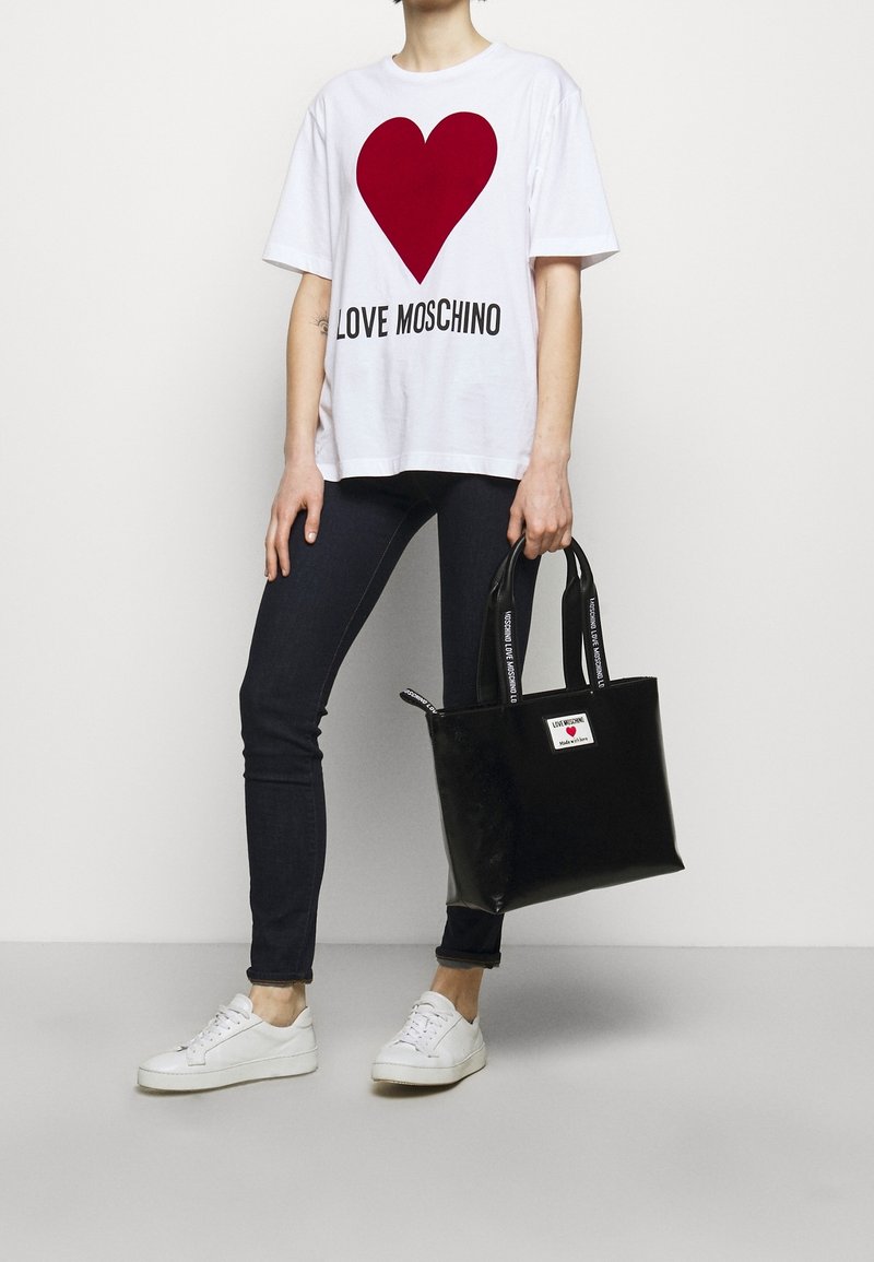 Weißes T-Shirt mit einem roten Herz-Design und dem Text "LOVE MOSCHINO", kombiniert mit dunklen Jeans und einer eleganten schwarzen Lackhandtasche. Weiße Sneakers.