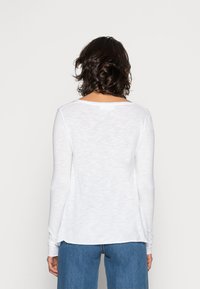 Ilga rankovė balta palaidinė, pagaminta iš minkštos, lengvos medžiagos, su laisvu siluetu ir subtilia tekstūra. Ypatybės: žemas nugaros iškirpimas ir šoninės skeltės.